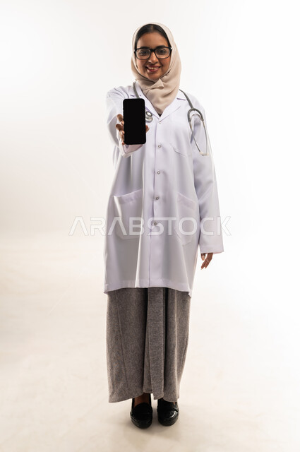 عرض شاشة سوداء فارغة، العمل في المراكز والمستشفيات، الطب والرعاية الصحية، بورتريه لطبيبة عربية خليجية عمانية محجبة ترتدي المعطف الطبي الأبيض تضع سماعة الفحص الطبية تحمل بيدها الهاتف المحمول, شابة سعودية تقوم بتقديم المساعدة الطبية، صورة لكامل الجسم، خلفية بيضاء