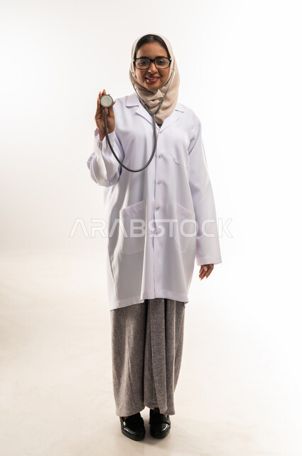 العمل في المراكز الصحية والمستشفيات، مفهوم الأنوثة والنعومة, الطب والرعاية الصحية، بورتريه لطبيبة عربية خليجية عمانية محجبة ترتدي المعطف الطبي الأبيض وترفع بيدها سماعة الفحص الطبية, شابة سعودية تقوم بتقديم المساعدة الطبية، صورة لكامل الجسم، خلفية بيضاء