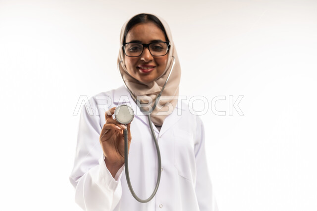 العمل في المراكز الصحية والمستشفيات، مفهوم الأنوثة والنعومة, الطب والرعاية الصحية، بورتريه مقرب لطبيبة عربية خليجية عمانية محجبة ترتدي المعطف الطبي الأبيض وترفع بيدها سماعة الفحص الطبية, شابة سعودية تقوم بتقديم المساعدة الطبية، خلفية بيضاء