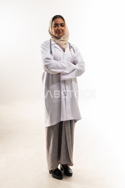 Providing medical assistance to patients, working in medical centers and hospitals, portrait of an Arab Gulf Omani female doctor wearing a white medical coat and an examination stethoscope standing with crossed hands, concept of medicine and health care, young Saudi woman looking at the camera with gestures of pleasure, full body image, white background