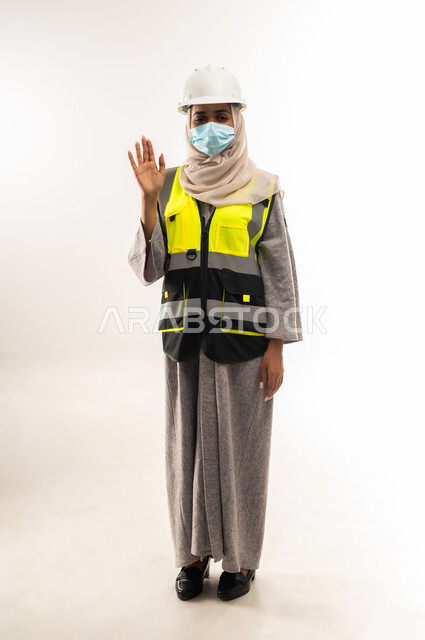 إدارة المشاريع الهندسية، مفهوم الوقاية من فايروس كورونا، وظائف ومهن نسائية, بورتريه لمهندسة معمارية عربية خليجية عمانية ترتدي خوذة وسترة الحماية وكمامة طبية ترفع يدها بايماءات القاء التحية والسلام، شابة سعودية تعمل في القطاع الهندسي، صورة لكامل الجسم، خلفية بيضاء