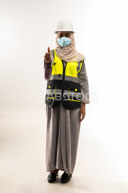 مفهوم الوقاية من فايروس كورونا، وظائف ومهن نسائية, إدارة المشاريع الهندسية، بورتريه لمهندسة معمارية عربية خليجية عمانية ترتدي خوذة وسترة الحماية والكمامة الطبية ترفع اصبع السبابة بايماءات الاعجاب، شابة سعودية تعمل في القطاع الهندسي، صورة لكامل الجسم، خلفية بيضاء