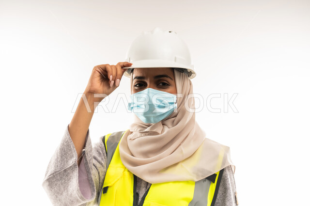 إدارة المشاريع الهندسية، مفهوم الوقاية من فايروس كورونا، وظائف ومهن نسائية, بورتريه لمهندسة معمارية عربية خليجية عمانية ترتدي خوذة وسترة الحماية والكمامة الطبية تنظر الى الكاميرا بايماءات الثقة بالنفس، شابة سعودية تعمل في القطاع الهندسي، خلفية بيضاء