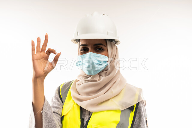 وظائف ومهن نسائية, مفهوم الوقاية من فايروس كورونا، إدارة المشاريع الهندسية، بورتريه مقرب لمهندسة معمارية عربية خليجية عمانية ترتدي خوذة وسترة الحماية والكمامة الطبية ترفع يدها بايماءات الاعجاب والرضا، شابة سعودية تعمل في القطاع الهندسي، خلفية بيضاء
