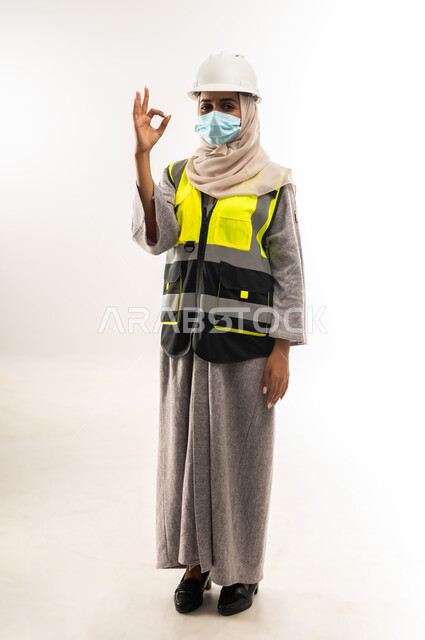 Women's jobs and professions, the concept of prevention from the Corona virus, engineering project management, a portrait of an Arab Gulf Omani female architect wearing a helmet, a protective vest and a medical mask, raising her hand with gestures of admiration and satisfaction, a young Saudi woman working in the engineering sector, a full body image, white background