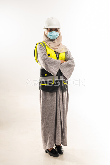 إدارة المشاريع الهندسية، مفهوم الوقاية من فايروس كورونا، وظائف ومهن نسائية, بورتريه لمهندسة معمارية عربية خليجية عمانية ترتدي خوذة وسترة الحماية والكمامة الطبية تقف بايدي متقاطعة، شابة سعودية تعمل في القطاع الهندسي، صورة لكامل الجسم، خلفية بيضاء