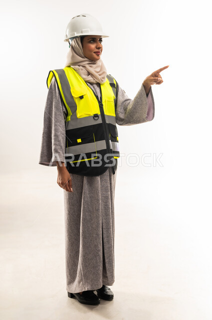 Locating a specific thing, engineering project management concept, women's jobs and careers, portrait of an Arab Gulf Omani female architect wearing a helmet and a protective vest pointing to a place, a young Saudi woman working in the engineering sector, full body image, white background