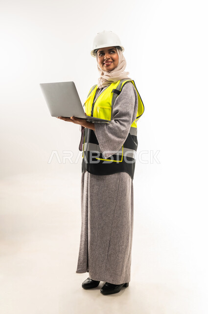 انجاز مهام العمل عبر الحاسوب المحمول, ادارة المشاريع الهندسية, بورتريه من الجانب لمهندسة عربية خليجية عمانية ترتدي سترة وخوذة الحماية تمسك الابتوب, العمل في القطاع الهندسي, شابة سعودية تقوم بدراسة أساسيات المشروع, صورة لكامل الجسم، خلفية بيضاء