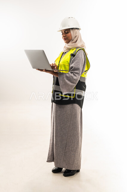ادارة المشاريع الهندسية, انجاز مهام العمل عبر الحاسوب المحمول, بورتريه من الجانب لمهندسة عربية خليجية عمانية ترتدي سترة وخوذة الحماية تمسك الابتوب, العمل في القطاع الهندسي, شابة سعودية تقوم بدراسة أساسيات المشروع, صورة لكامل الجسم، خلفية بيضاء