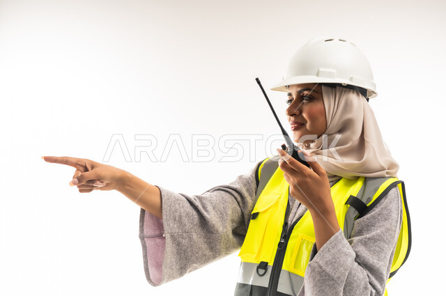 تحديد موقع شيء معين، مهن وظائف هندسية، متابعة سير الاعمال، العمل في القطاع الهندسي، الإشراف على المشاريع، بورتريه مقرب لمهندسة عربية خليجية عمانية ترتدي سترة وخوذة الحماية تقوم باستخدام جهاز الاتصال اللاسلكي، شابة سعودية تشير باصبع السبابة لمكان ما، خلفية بيضاء