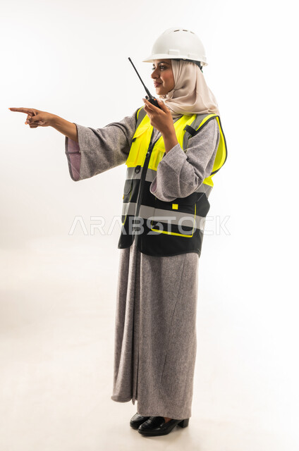 Engineering jobs, business follow-up, working in the engineering sector, project supervision, side portrait of an Arab Gulf Omani female engineer wearing a protective jacket and helmet using a walkie-talkie, a young Saudi woman pointing her index finger somewhere, full body image, white background