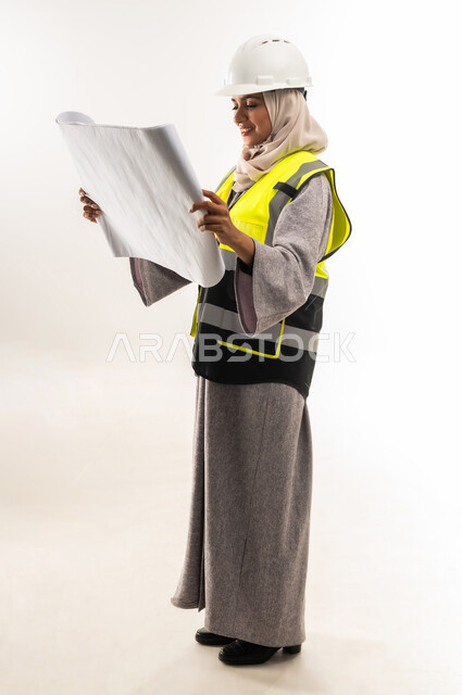 مهن ووظائف نسائية هندسية، العمل في القطاع الهندسي، مخططات و مشاريع اعمارية، بورتريه من الجانب لامرأة عربية خليجية عمانية محجبة ترتدي خوذة وسترة الحماية تحمل بيدها مخطط المشروع، شابة سعودية تقوم بالاشراف على العمل، صورة لكامل الجسم، خلفية بيضاء