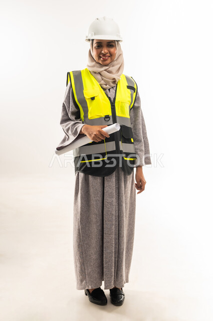 العمل في القطاع الهندسي، مخططات و مشاريع اعمارية، مهن ووظائف نسائية هندسية، بورتريه لامرأة عربية خليجية عمانية محجبة ترتدي خوذة وسترة الحماية تحمل بيدها مخطط المشروع، شابة سعودية تقوم بالاشراف على العمل، صورة لكامل الجسم، خلفية بيضاء