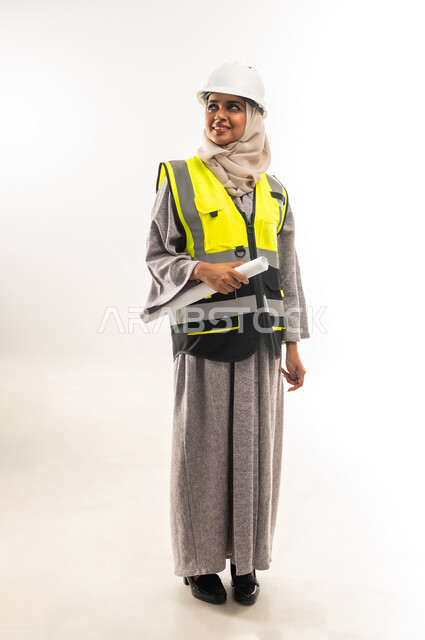 مخططات و مشاريع اعمارية، العمل في القطاع الهندسي، مهن ووظائف نسائية هندسية، بورتريه لامرأة عربية خليجية عمانية محجبة ترتدي خوذة وسترة الحماية تحمل بيدها مخطط المشروع، شابة سعودية تقوم بالاشراف على العمل، صورة لكامل الجسم، خلفية بيضاء