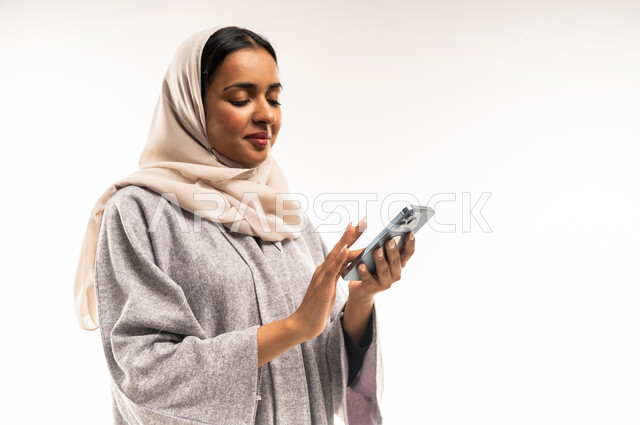 إيماءات السعادة والسرور، الدردشة مع الأهل والأصدقاء، استخدام الاجهزة التقنية الحديثة، بورتريه مقرب لامرأة عربية خليجية عمانية محجبة تحمل بيدها الهاتف المحمول، شابة سعودية تقوم بتصفح وسائل التواصل الاجتماعي، خلفية بيضاء