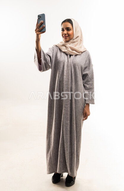 التقاط صورة سيلفي تذكارية، استخدام جهاز تقني حديث ومتطور، بورتريه لامرأة عربية خليجية عمانية محجبة ترتدي عباءة تضع نظارة طبية تقوم بإجراء مكالمة فيديو، شابة سعودية تستخدام الهاتف لمحمول، صورة لكامل الجسم، خلفية بيضاء