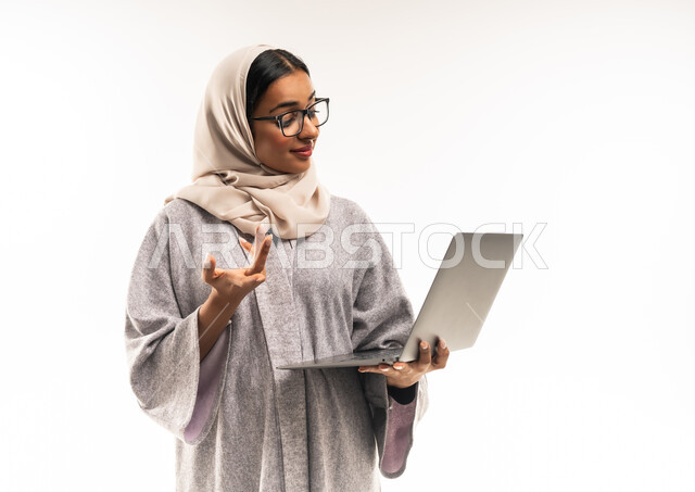 استخدام الأجهزه التقنية الحديثة, اجراء مكالمة فيديو, مفهوم التعليم عن بعد, بورتريه مقرب لامرأة عربية خليجية عمانية محجبة تضع نظارة طبية تحمل اللابتوب ترفع يدها بايماءات السؤال، شابة سعودية تقوم بمتابعة المحاضرات الجامعية عبر منصات التعليم المتطورة، خلفية بيضاء