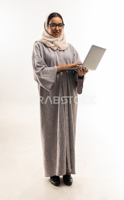 إدارة شؤون الأعمال وتنظيمه, التواصل مع الأهل والأصدقاء عن بعد، استخدام التقنيات الحديثة، بورتريه لامراة عربية خليجية عمانية محجبة ترتدي العباءة تضع نظارة طبية تحمل بيدها الحاسوب المحمول, شابة سعودية تقوم بالنقر على لوحة المفاتيح، صورة الجسم بالكامل, خلفية بيضاء