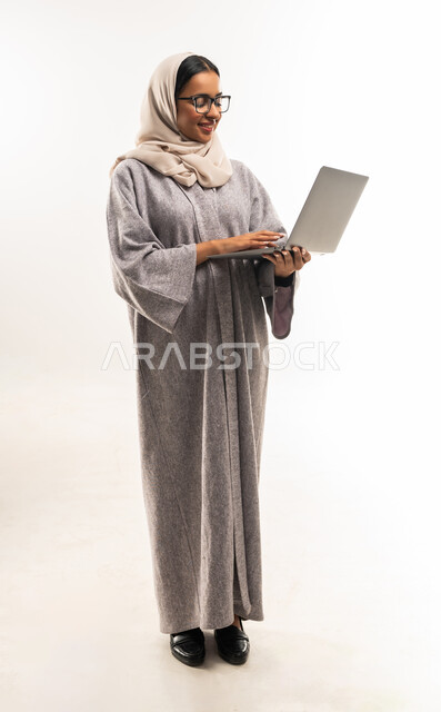 Communicating with family and friends remotely, managing and organizing business affairs, using modern technologies, portrait of a veiled Gulf Arab Omani woman wearing an abaya, wearing medical glasses and holding a laptop in her hand, a young Saudi woman clicking on the keyboard, full body image, white background