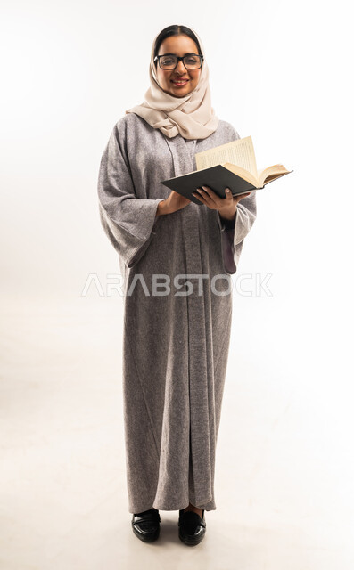Self-learning culture, spending time learning and educating yourself, enjoying reading books, stories and novels, portrait of a veiled Gulf Omani Arab woman wearing an abaya, wearing glasses and holding a book in her hands, a young Saudi woman working hard and studying, full body image, white background