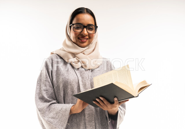 الاستمتاع بقراءة الكتب والقصص والروايات، قضاء الوقت بالتعلم والتثقف، بورتريه مقرب لامرأة عربية خليجية عمانية محجبة ترتدي العباءة تضع النظارة الطبية تحمل بيديها كتاب، شابة سعودية تقوم الاجتهاد والدراسة، ثقافة التعلم الذاتي، خلفية بيضاء