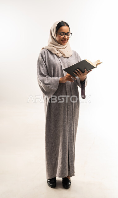 Spending time learning and educating yourself, enjoying reading books, stories and novels, portrait of a veiled Gulf Arab Omani woman wearing an abaya, wearing glasses and holding a book in her hands, a young Saudi woman working hard and studying, self-learning culture, full body image, white background