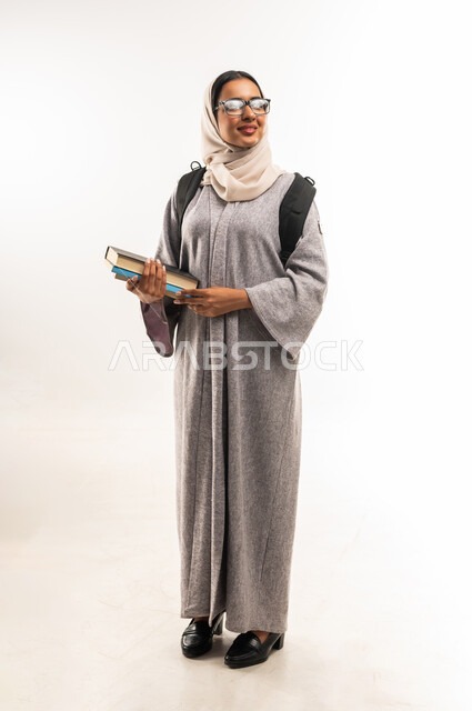 مفهوم التعليم الأكاديمي، الاستعداد للعام الدراسي الجديد, بورتريه لطالبة جامعية عربية خليجية عمانية مبتسمة ترتدي العباءة والنظارة الطبية تحمل الحقيبة تمسك بيدها مجموعة من الكتب, شابة سعودية تنظر الى مكان ما بايماءات السعادة والسرور, صورة لكامل الجسم، خلفية بيضاء