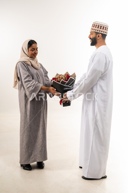 زوجان سعيدان يحتفلان بعيد الحب , بورتريه لرجل عربي خليجي عماني يرتدي الدشداشة و الكمة يقدم لزوجته باقة من الورد الاحمر، إيماءات الدهشة والفرح، الاحتفال بالمناسبات الرومنسية، حفلة ذكرى الزواج، الجسم بالطول الكامل، خلفية بيضاء