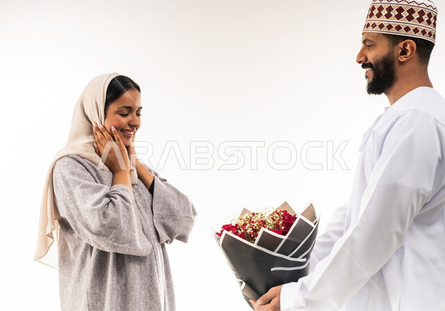 تقديم الزهور في الأعياد والمناسبات السعيدة، حفلة ذكرى الزواج، زوجان سعيدان يحتفلان بعيد الحب , بورتريه مقرب لرجل عربي خليجي عماني يرتدي الدشداشة و الكمة يقدم لزوجته باقة من الورد الاحمر، إيماءات الدهشة والفرح، انتقاء باقة مميزة من الورود، خلفية بيضاء