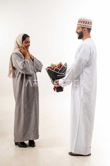 الاحتفال بالمناسبات الرومنسية، زوجان سعيدان يحتفلان بعيد الحب , بورتريه لرجل عربي خليجي عماني يرتدي الدشداشة و الكمة يقدم لزوجته باقة من الورد الاحمر، إيماءات الدهشة والفرح، حفلة ذكرى الزواج، الجسم بالطول الكامل، خلفية بيضاء