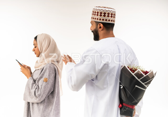 الاحتفال بالمناسبات الرومنسية، حفلة ذكرى الزواج، زوجان سعيدان يحتفلان بعيد الحب , بورتريه مقرب لرجل عربي خليجي عماني يرتدي الدشداشة و الكمة يقدم لزوجته باقة من الورد الاحمر، إيماءات الدهشة والفرح، امراة عمانية تقوم باستخدام الهاتف المحمول، خلفية بيضاء