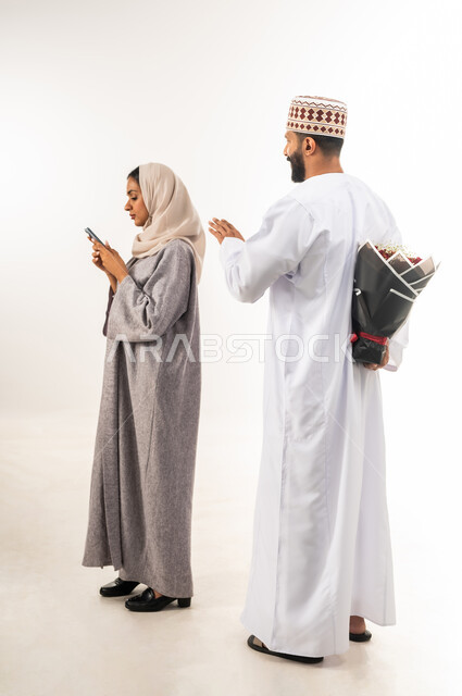 Exchange of affection between family members, wedding anniversary party, happy couple celebrating Valentine's Day, portrait of an Arab Gulf Omani man wearing a dishdasha and a kummah offering his wife a bouquet of red roses, gestures of surprise and joy, celebrating romantic occasions, full body, white background