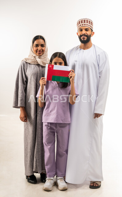 Celebrating Oman National Day, participating in national holidays and occasions, pride in Omani symbols and flag, portrait of an Arab Gulf Omani girl wearing and holding the flag of the Sultanate of Oman in her hand, standing with her parents, the concept of love of the homeland and belonging to it, full body, white background