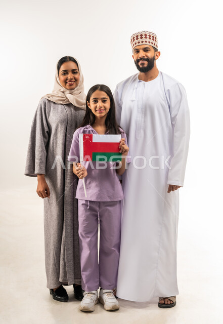 المشاركة بالاعياد والمناسبات الوطنية، الاحتفال بذكرى اليوم الوطني العماني، الاعتزاز والفخر بالرموز والراية العمانية، بورتريه لفتاة عربية خليجية عمانية يرتدي تحمل بيدها علم سلطنة عمان تقف مع والديها، مفهوم حب الوطن والإنتماء إليه، الجسم بالطول الكامل، خلفية بيضاء