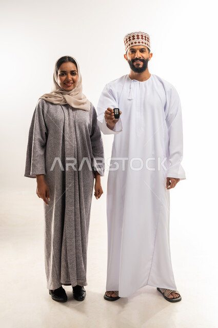 مشاركة الأسرة فرحة تملك شيء جديد، ايماءات السعادة والسرور لإقتناء سيارة جديدة، الحفاظ على الاستقرار والأمان الأسري، بورتريه لرجل عربي خليجي عماني يرتدي الدشداشة والكمة يقف برفقة زوجته يحمل بيده مفتاح التحكم الالكتروني، الطول الكامل للجسم، خلفية بيضاء