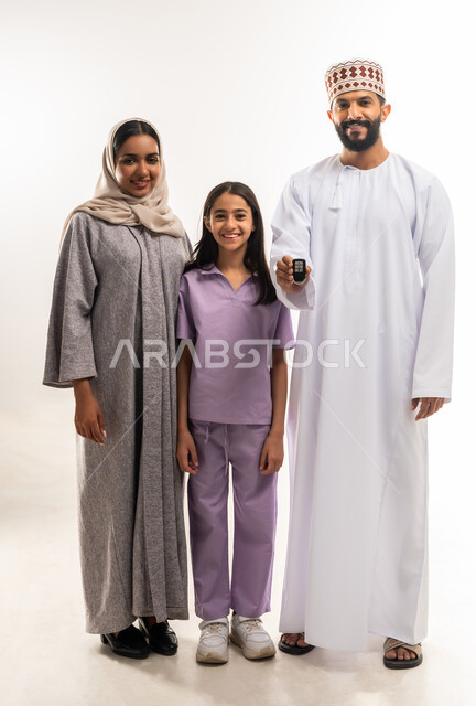 ايماءات السعادة والسرور لإقتناء سيارة جديدة، الحفاظ على الاستقرار والأمان الأسري، بورتريه لرجل عربي خليجي عماني يرتدي الدشداشة والكمة يقف برفقة عائلته يحمل بيده مفتاح التحكم الالكتروني، مشاركة الأسرة فرحة تملك شيء جديد، الطول الكامل للجسم، خلفية بيضاء
