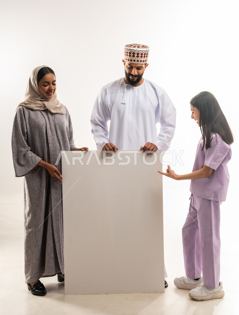 الاعلانات والعروض التسويقية، استخدام لوحات توضيحية ووسائل تعليمية، بورتريه لرجل عربي خليجي عماني مبتسم يرتدي الدشداشه والكمه يقف مع زوجته وابنته وبجانبهم لوح ابيض كبير،  موك اب فارغ، خلفية بيضاء