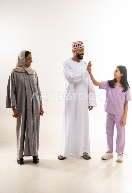 Exchanging entertaining dialogues and discussions, an Omani family having a good time, a portrait of an Arab Gulf Omani man wearing a dishdasha and a kummah standing with his wife and daughter with gestures of happiness and pleasure, strengthening social relations, full-length body, white background