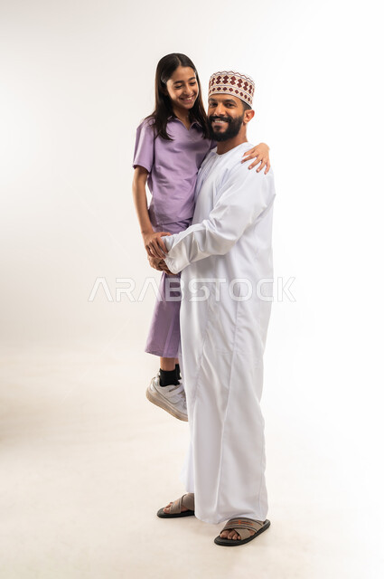عائلة عمانية تقضي اوقات ممتعة ومسلية، الاهتمام بالأبناء ورعايتهم، تعزيز العلاقات الاجتماعية، بورتريه لرجل عربي خليجي عماني يرتدي الدشداشة والكمة يحمل ابنته بايماءات السعادة والسرور، الجسم بالطول الكامل، خلفية بيضاء