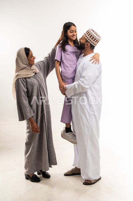 الاهتمام بالأبناء ورعايتهم، عائلة عمانية تقضي اوقات ممتعة ومسلية، بورتريه لرجل عربي خليجي عماني يرتدي الدشداشة والكمة يحمل ابنته ويقف مع زوجته بايماءات السعادة والسرور، تعزيز العلاقات الاجتماعية، الجسم بالطول الكامل، خلفية بيضاء