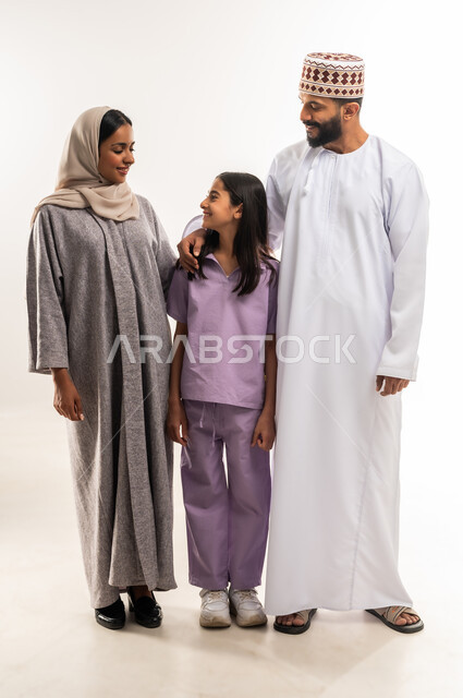تبادل الحوارات والنقاشات المسلية، عائلة عمانية تقضي اوقات ممتعة، الرعايه والاهتمام بالأبناء، بورتريه لرجل عربي خليجي عماني يرتدي الدشداشة والكمة يقف مع زوجته وابنته بايماءات السعادة والسرور، تعزيز العلاقات الاجتماعية، الجسم بالطول الكامل، خلفية بيضاء