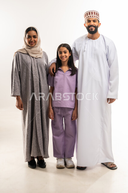 عائلة عمانية تقضي اوقات ممتعة، النظر إلى الكاميرا بايماءات السعادة والسرور، الرعايه والاهتمام بالأبناء، بورتريه لرجل عربي خليجي عماني يرتدي الدشداشة والكمة يقف مع زوجته وابنته، تعزيز العلاقات الاجتماعية، الجسم بالطول الكامل، خلفية بيضاء