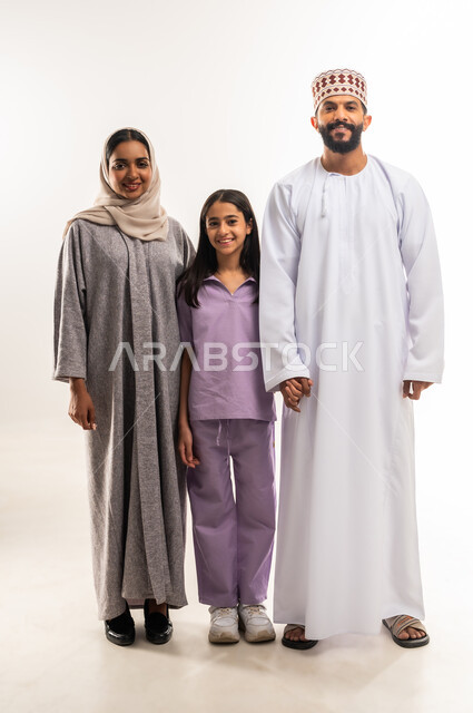 النظر إلى الكاميرا بايماءات السعادة والسرور، عائلة عمانية تقضي اوقات ممتعة، تبادل الحوارات والنقاشات المسلية، بورتريه لرجل عربي خليجي عماني يرتدي الدشداشة والكمة يقف مع زوجته وابنته، تعزيز العلاقات الاجتماعية، الجسم بالطول الكامل، خلفية بيضاء