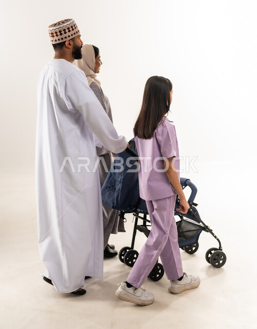 The importance of parents and their essential role in raising and educating children, caring for and paying attention to children, portrait of an Arab Gulf Omani man wearing a dishdasha and a kummah standing with his wife and daughter and pulling a baby carriage, going on family outings and tours, an Omani family spending enjoyable times, white background