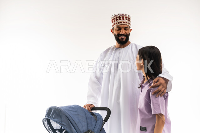 Going on family outings and tours, taking care of children, the importance of the father and his essential role in raising and educating children, a portrait of an Arab Gulf Omani man wearing a dishdasha and a kummah pulling a baby carriage with his daughter, an Omani family spending fun and entertaining times, white background