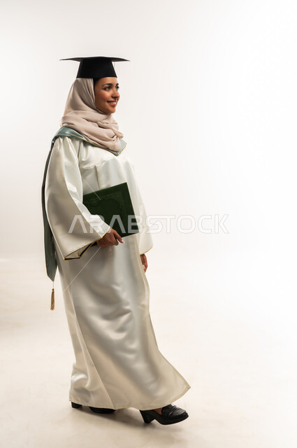 الشعور بالفخر والثقة بالنفس، الاحتفال بإنهاء الدراسة الجامعية, بورتريه لخريجة عربية خليجية عمانية محجبة مبتسمة ترتدي العباءة والقبعة تمسك بيدها وثيقة التخرج، شابة سعودية تنظر بإيماءات السعادة بالوصول لحلم النجاح، الجسم الكامل بالطول، خلفية بيضاء