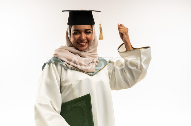 التفوق والتخرج من الجامعة، الاحتفال بإنهاء الدراسة الجامعية, بورتريه مقرب لخريجة عربية خليجية عمانية محجبة مبتسمة ترتدي العباءة والقبعة تمسك بيدها وثيقة التخرج، شابة سعودية ترفع قبضة يدها بإيماءات السعادة بالوصول لحلم النجاح، خلفية بيضاء