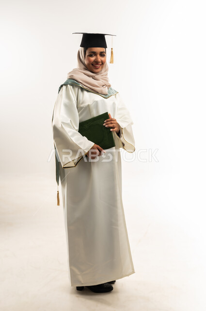 Excellence and graduation from university, celebration of completing university studies, portrait of a smiling veiled Gulf Arab Omani graduate wearing an abaya and a hat holding her graduation document, a young Saudi woman looking with gestures of happiness at reaching the dream of success, full body length, white background