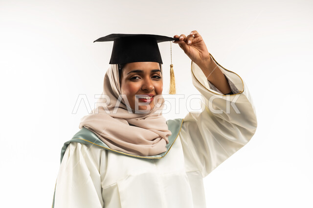 تعابير النجاح والفوز، الاحتفال بإنهاء الدراسة الجامعية, بورتريه مقرب لخريجة عربية خليجية عمانية محجبة مبتسمة ترتدي العباءة وتمسك قبعة التخرج، شابة سعودية تنظر بإيماءات السعادة بالوصول لحلم النجاح، خلفية بيضاء