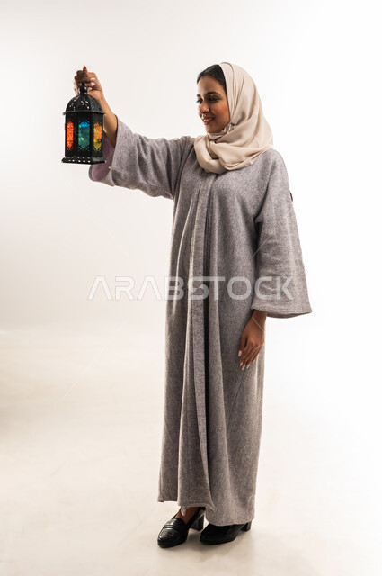 الاستعداد لاستقبال شهر رمضان المبارك, فوانيس وزينة الأعياد والمناسبات الاسلامية, شابة سعودية تنظر إلى المصباح بإيماءات السرور, بورتريه لامرأة عربية خليجية عمانية محجبة ترتدي العباءة تمسك بيديها فانوس ملون, الجسم بالطول الكامل، خلفية بيضاء